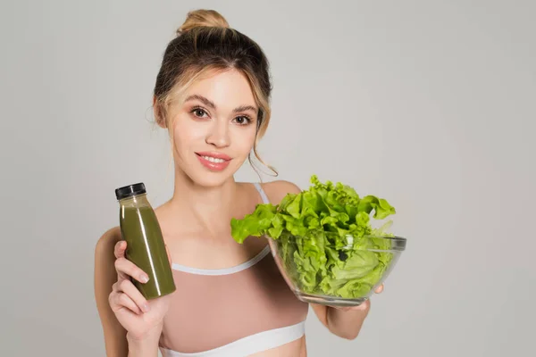 Mujer feliz con la piel perfecta sosteniendo batido fresco y tazón de lechuga aislado en gris - foto de stock