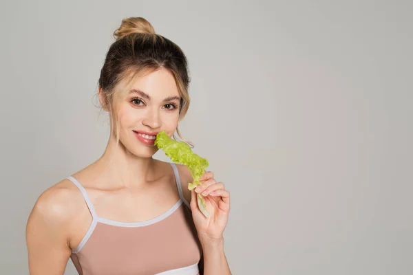 Pretty woman with natural makeup holding fresh lettuce and looking at camera isolated on grey — Stock Photo