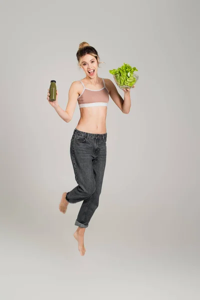 Excité pieds nus femme avec mince corps lévitant avec laitue et bouteille de smoothie sur fond gris — Photo de stock