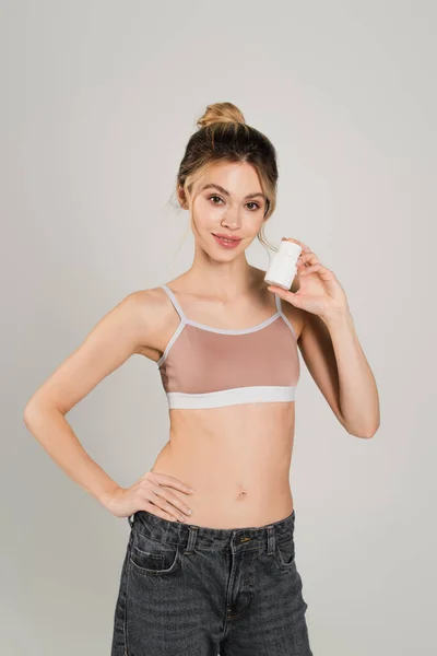 Mujer complacida en la parte superior de los deportes de pie con la mano en la cadera y mostrando vitaminas aisladas en gris — Stock Photo