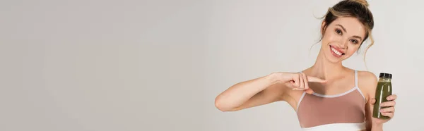 Mujer alegre en top deportivo apuntando a la botella de batido fresco y natural aislado en gris, bandera - foto de stock