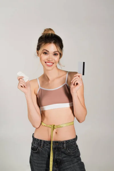 Mulher alegre com fita métrica na cintura segurando vitaminas e cartão de crédito isolado em cinza — Fotografia de Stock
