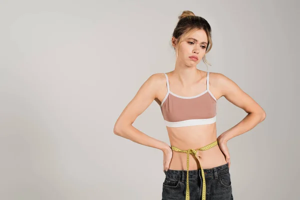 Upset woman in sports top standing with measuring tape on waist and hands on hips isolated on grey — Stock Photo