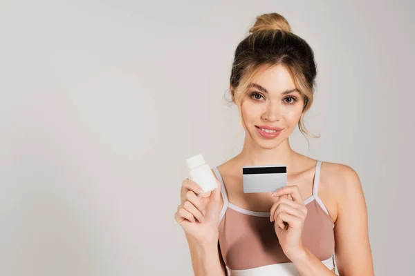 Jolie femme avec maquillage naturel et peau parfaite tenant carte de crédit et vitamines isolées sur gris — Photo de stock