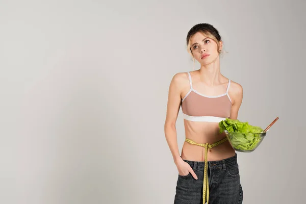 Upset woman with measuring tape on waist holding bowl of fresh lettuce isolated on grey — Stock Photo