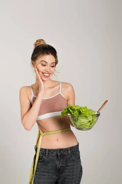 Amazed woman with measuring tape on waist looking at bowl with lettuce and touching face isolated on grey — Stock Photo