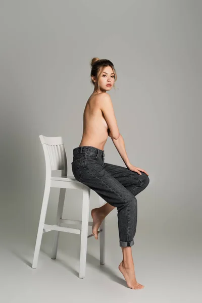 Piena lunghezza di mezza donna nuda a piedi nudi in jeans seduta su sedia bianca su sfondo grigio — Foto stock