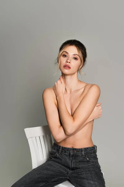 Pretty half naked woman covering breast with crossed arms while sitting on white chair isolated on grey — Stock Photo