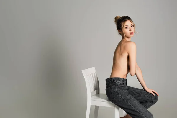 Giovane donna mezza nuda in jeans seduta sulla sedia bianca e guardando la fotocamera isolata sul grigio — Foto stock