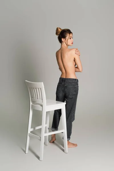 Rückansicht einer halbnackten Frau in Jeans, die in der Nähe eines weißen Stuhls steht und vor grauem Hintergrund wegschaut — Stockfoto