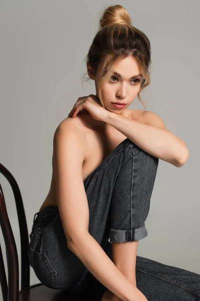 Young half naked woman in jeans sitting on wooden chair and looking away isolated on grey — Stock Photo