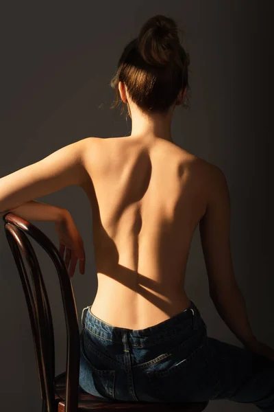 Vista posteriore di donna mezza nuda in jeans seduta su sedia di legno in luce isolata su grigio — Foto stock