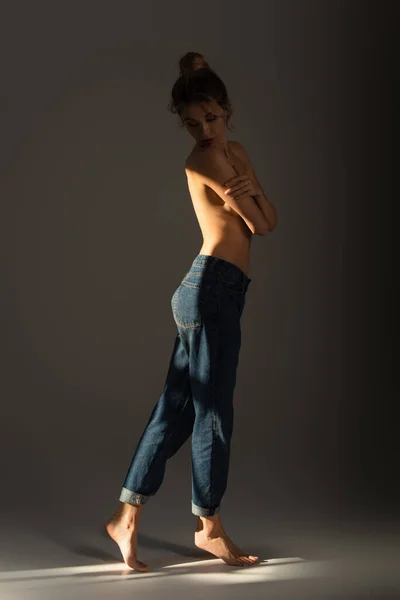 Full length of half nude barefoot woman in jeans covering bust with crossed arms on grey background with lighting — Stock Photo