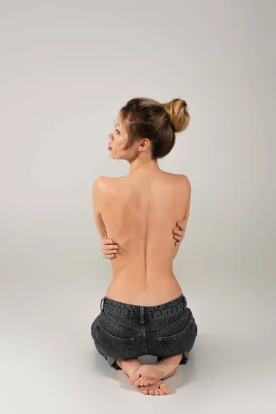 Vista posteriore di mezza donna nuda a piedi nudi in jeans seduta e guardando lontano su sfondo grigio — Foto stock