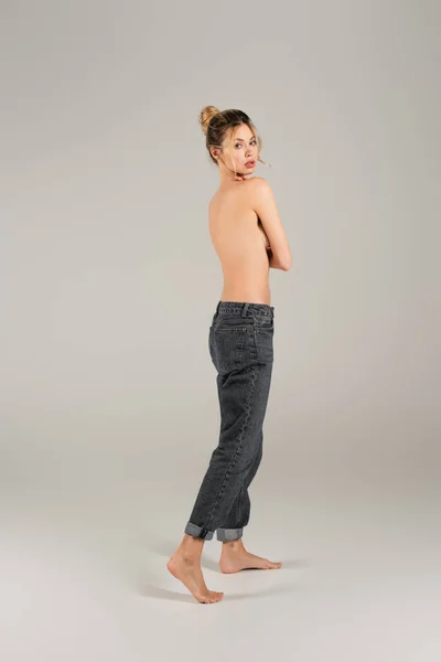 Piena lunghezza di sottile donna mezza nuda in jeans in piedi a piedi nudi e guardando la fotocamera su sfondo grigio — Foto stock