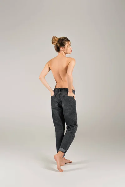 Back view de mulher descalça semi-nua de pé com as mãos nos bolsos traseiros de jeans no fundo cinza — Fotografia de Stock