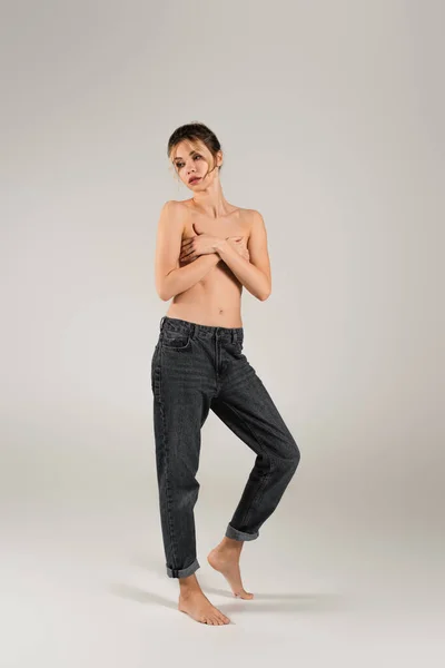 Full length of half naked barefoot woman in jeans covering breast with crossed arms and looking away on grey background — Stock Photo