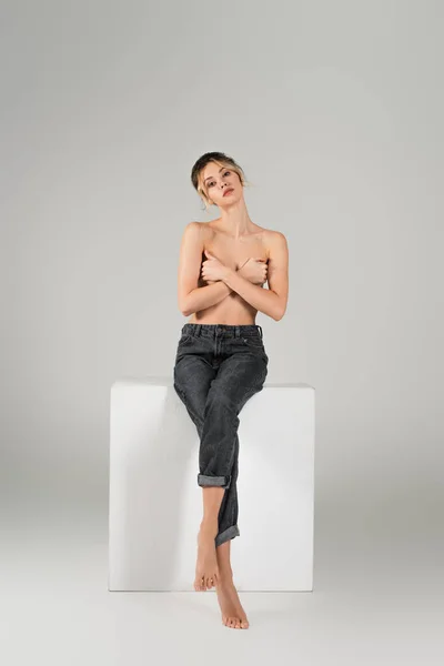 Full length of barefoot and half naked woman in jeans covering bust with crossed arms on white cube on grey background — Stock Photo