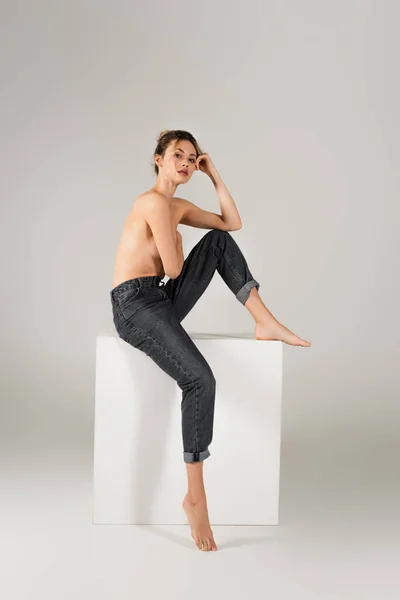 Sexy half nude and barefoot woman in jeans sitting on white cube and looking at camera on grey background — Stock Photo