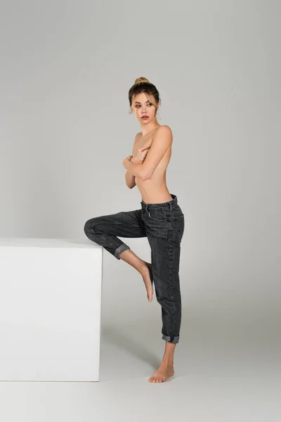 Half dressed barefoot woman in jeans posing near white cube and covering bust with crossed arms on grey background — Stock Photo