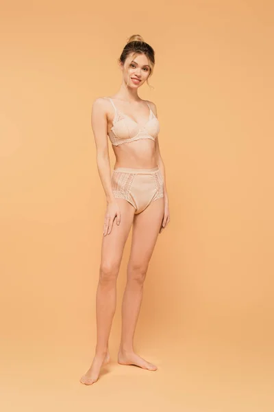 Longitud completa de la mujer en forma en ropa interior de encaje sonriendo a la cámara sobre fondo beige - foto de stock