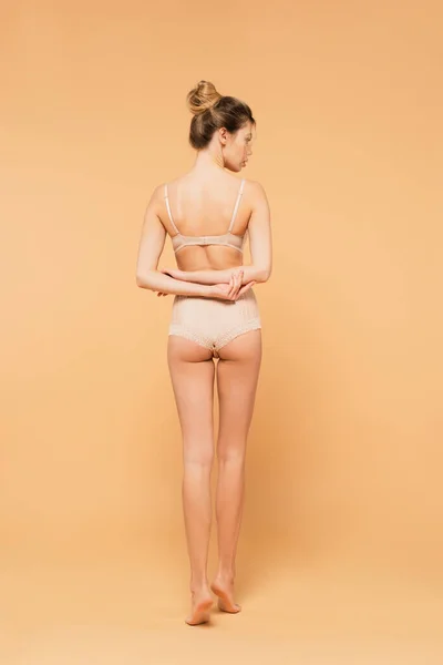Вид ззаду босоніж жінка з ідеальним тілом, що стоїть в нижній білизні з руками позаду на бежевому фоні — стокове фото