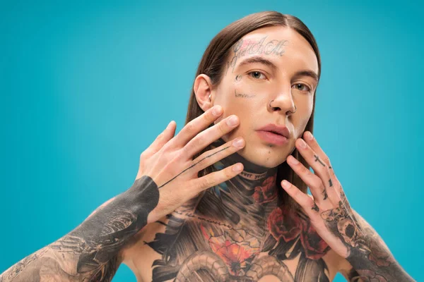 Young man with tattoos touching smooth face while looking at camera isolated on blue — Stock Photo