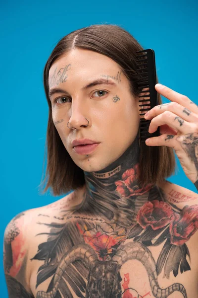 Young and pierced man with tattoos brushing hair with comb isolated on blue — Stock Photo