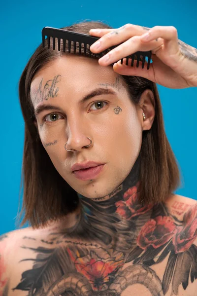 Hombre joven y tatuado cepillando el cabello con peine aislado en azul - foto de stock