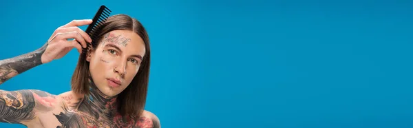 Young and pierced man with tattoos brushing hair with comb isolated on blue, banner — Stock Photo