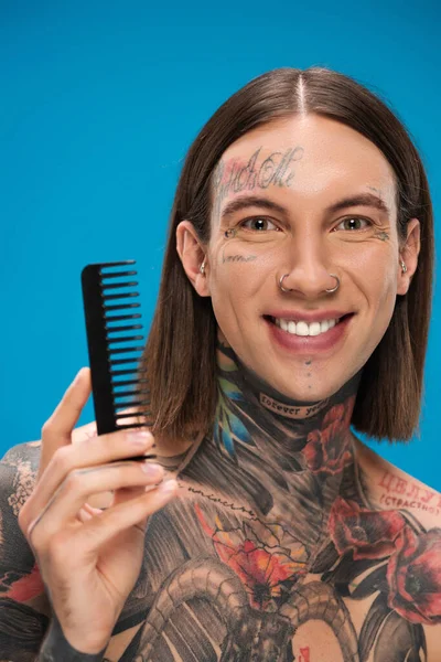 Happy and pierced young man with tattoos holding comb isolated on blue — Stock Photo