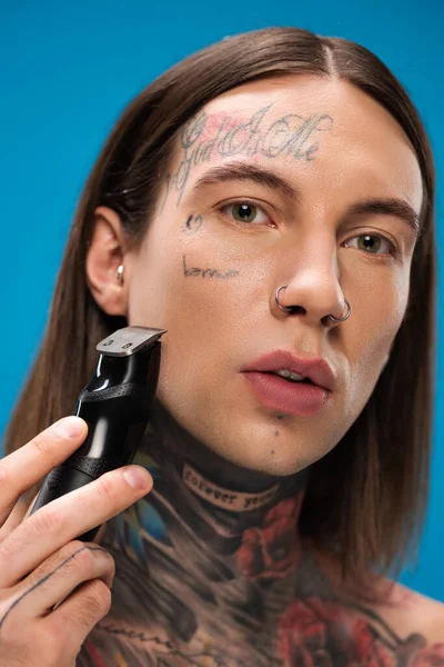 Close up de homem jovem e perfurado com tatuagens de barbear com lâmina elétrica isolada em azul — Fotografia de Stock