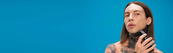 Молодой и пирсинг мужчина с татуировками бритья с электрической бритвой изолированы на синий, баннер — стоковое фото