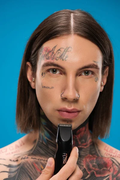 Hombre joven y perforado con tatuajes recortar la cara con afeitadora eléctrica aislado en azul - foto de stock
