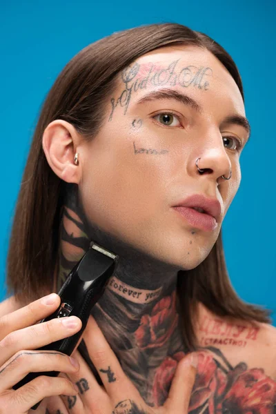 Young and tattooed man shaving face with electric razor isolated on blue — Stock Photo