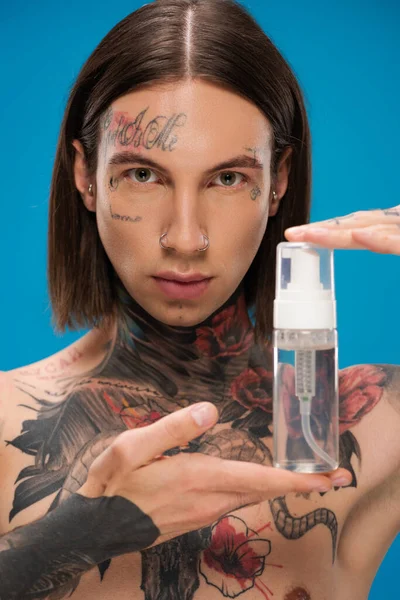 Junger tätowierter Mann hält Flasche mit Reinigungsschaum isoliert auf blau — Stockfoto