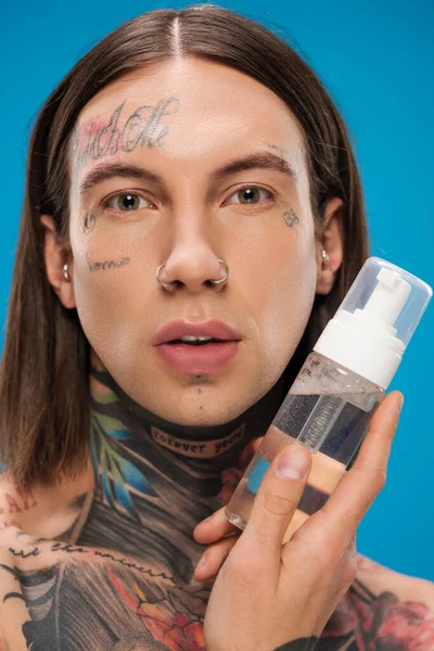 Tattooed young man holding bottle with cleansing foam isolated on blue — Stock Photo