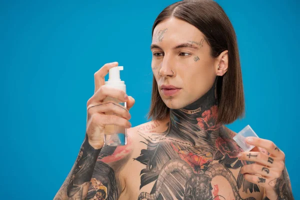 Jeune homme tatoué regardant bouteille avec mousse nettoyante isolé sur bleu — Photo de stock