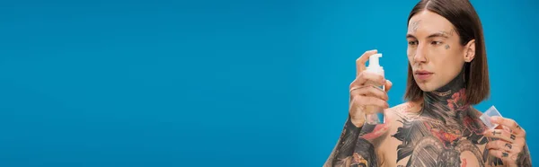Giovane uomo tatuato guardando bottiglia con schiuma detergente isolato su blu, banner — Foto stock