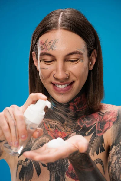 Sorridente e tatuado homem segurando garrafa com espuma de limpeza isolado em azul — Fotografia de Stock