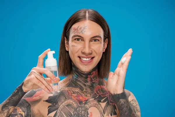 Hombre alegre y tatuado sosteniendo botella con espuma limpiadora aislada en azul - foto de stock