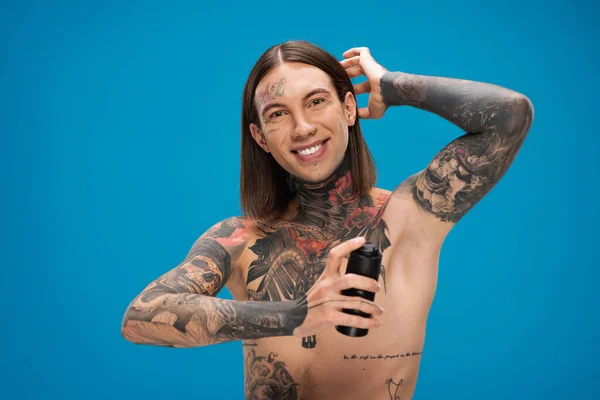 Joyeux et tatoué jeune homme pulvérisation déodorant isolé sur bleu — Photo de stock