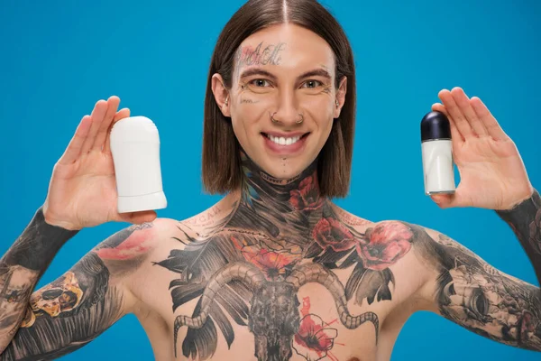 Hombre alegre y tatuado sosteniendo diferentes desodorantes aislados en azul - foto de stock
