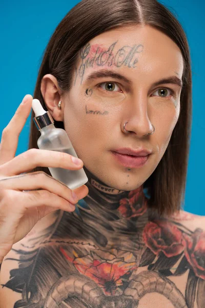 Young and pierced man with tattoos holding bottle with serum isolated on blue — Stock Photo