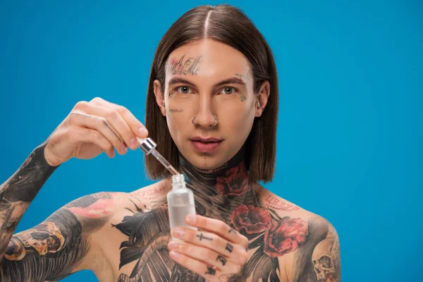 Jeune homme percé de tatouages tenant bouteille avec sérum hydratant isolé sur bleu — Photo de stock