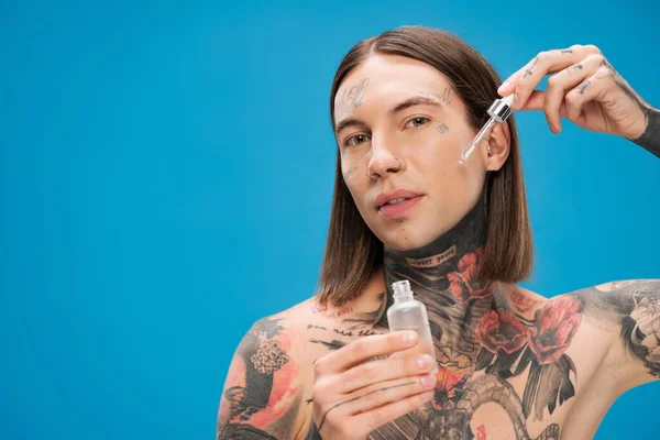 Giovane e tatuato bottiglia uomo tenendo mentre si applica siero idratante isolato su blu — Foto stock