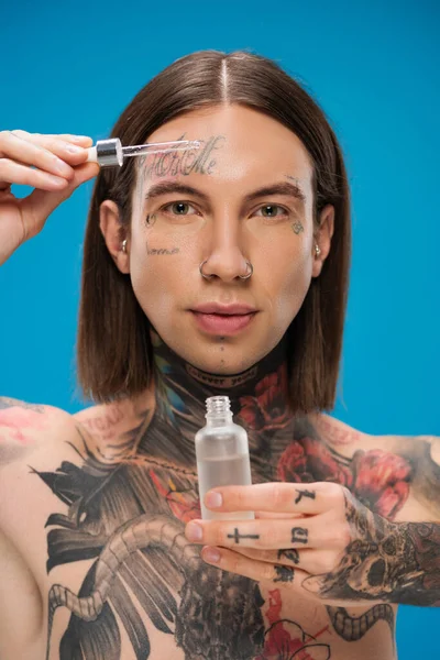 Jeune homme tatoué tenant bouteille et pipette avec sérum hydratant isolé sur bleu — Photo de stock