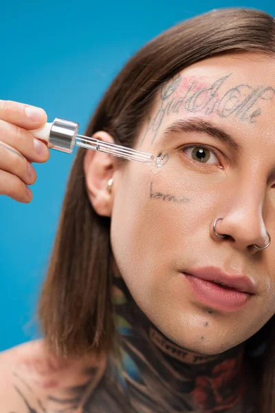 Крупным планом молодого татуированного мужчины, использующего увлажняющую сыворотку с пипеткой, выделенной на голубом — стоковое фото