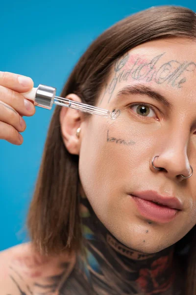 Nahaufnahme eines tätowierten jungen Mannes, der feuchtigkeitsspendendes Serum mit Pipette auf blauem Grund aufträgt — Stockfoto