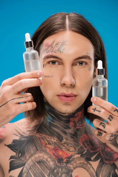 Hombre joven y tatuado sosteniendo botellas con suero hidratante aislado en azul - foto de stock
